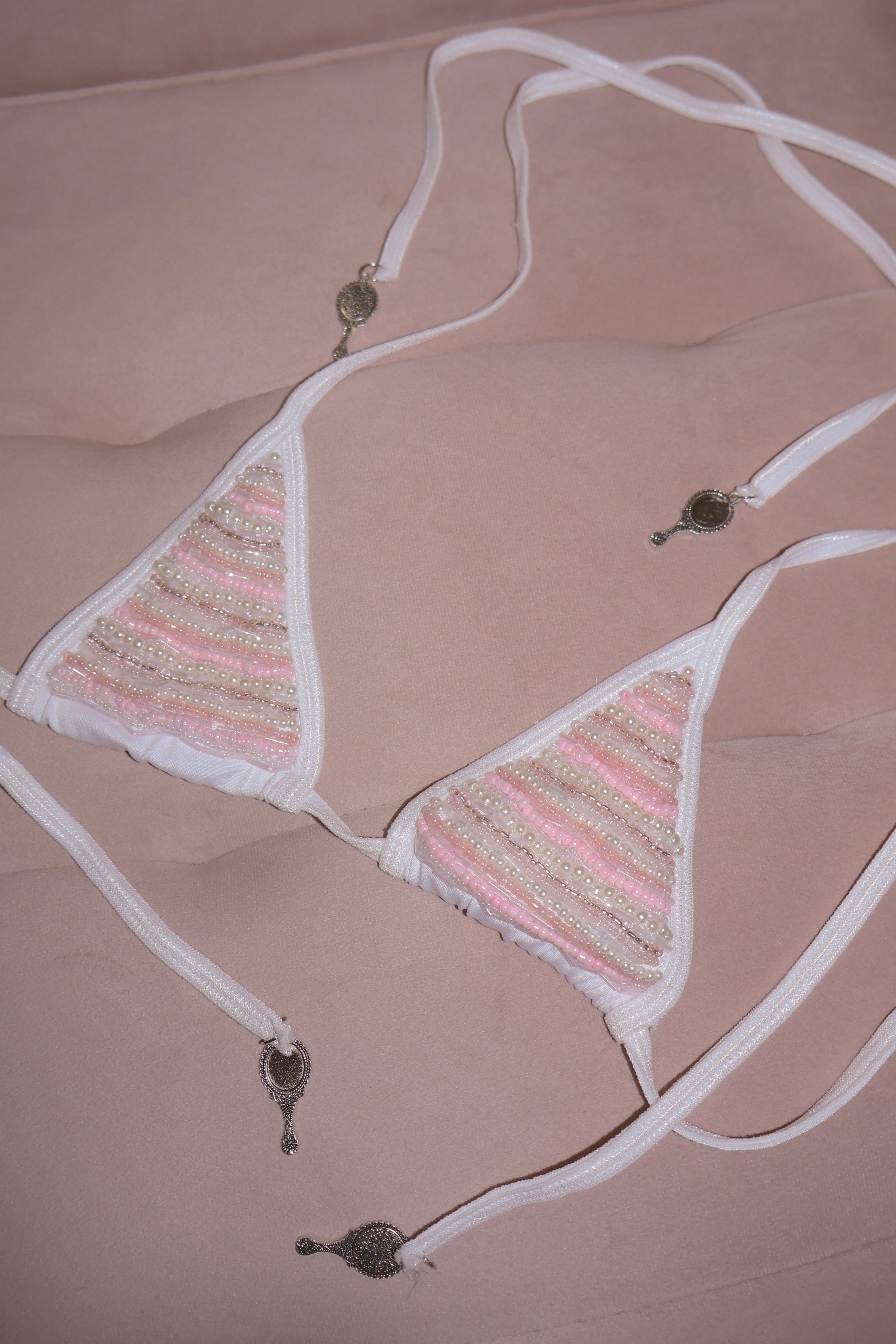 A triangle bikini top with light pink and beige striped woven fabric panels, white trim, and white adjustable ties with decorative metal charms, laid flat on a dusty pink fabric background.